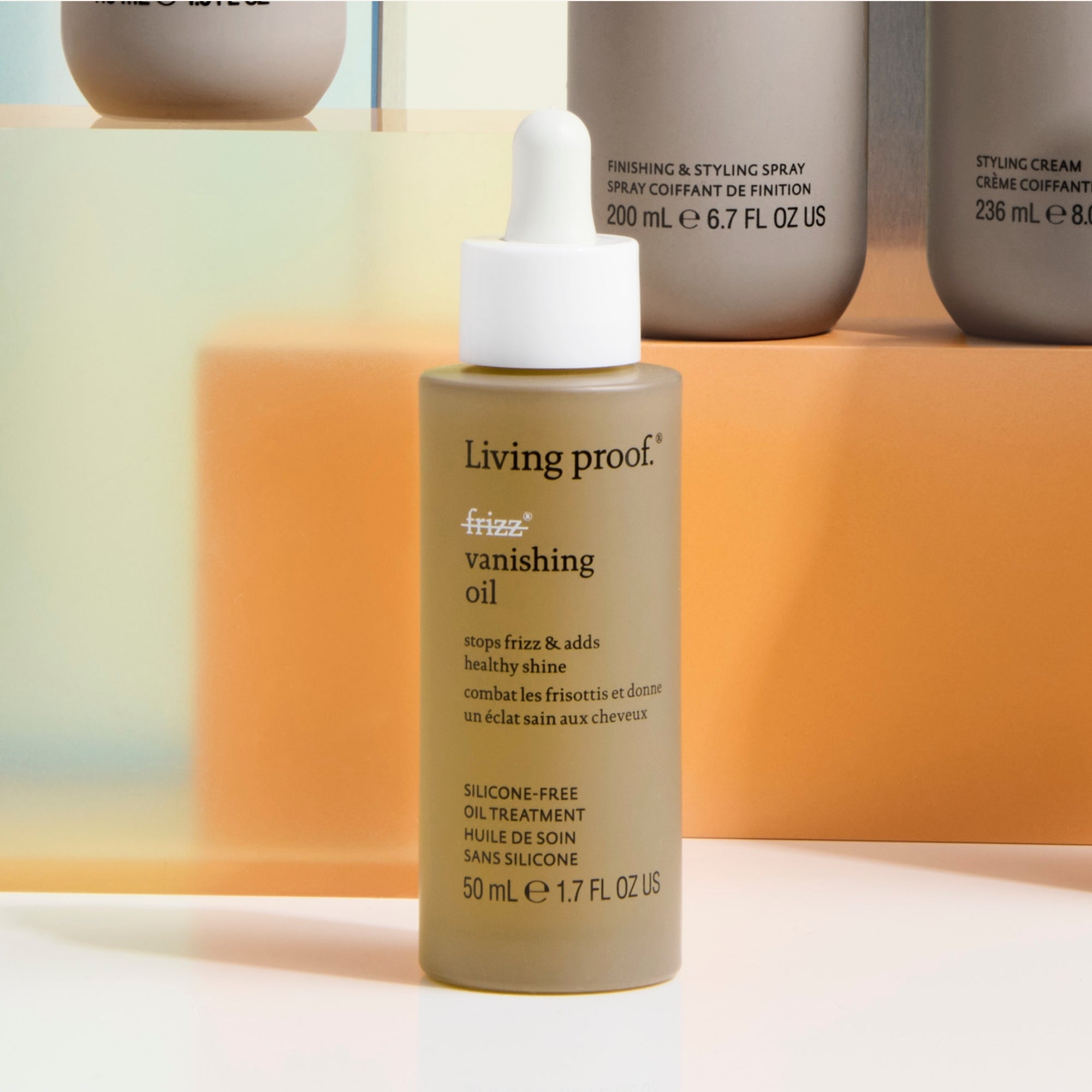 A bottle of No Frizz Vanishing Oil with a white dropper cap sits on a light surface, surrounded by other hair products. The label features its hydrating formula, humidity protection, and benefits for shiny hair.