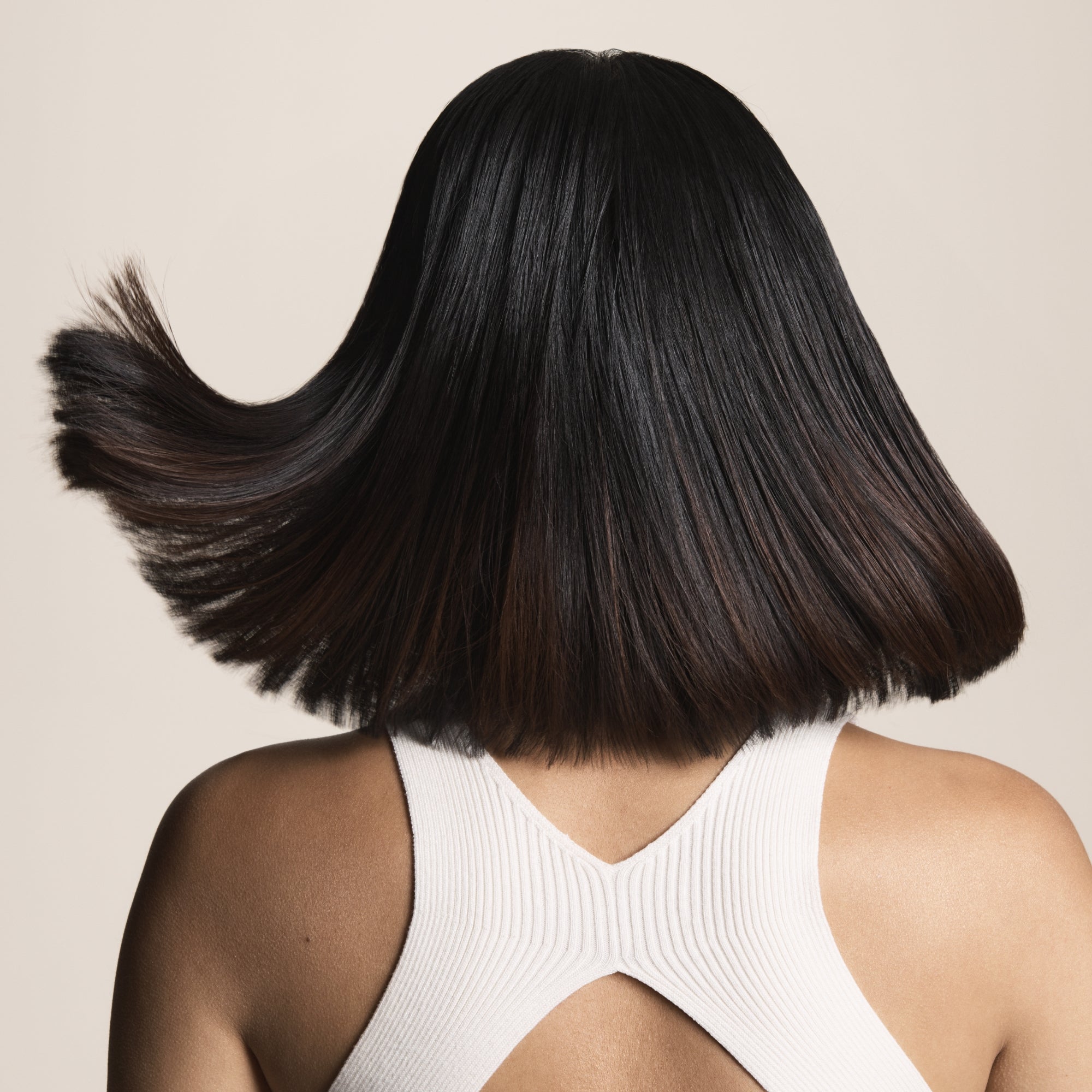 A person with straight, shoulder-length dark hair, seen from behind in a white sleeveless top with a back cutout, shows off shiny, swinging hair full of movement thanks to the Clarifying Detox Shampoo that removes buildup.