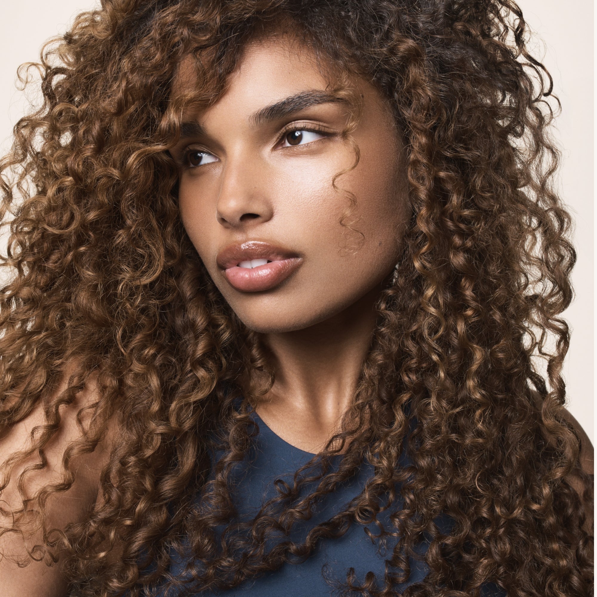 A woman with long, voluminous, curly brown hair—freshly washed with Clarifying Detox Shampoo—looks to the side. Her smooth skin, defined brows, and glossy lips complement her sleeveless dark blue top against a light background.
